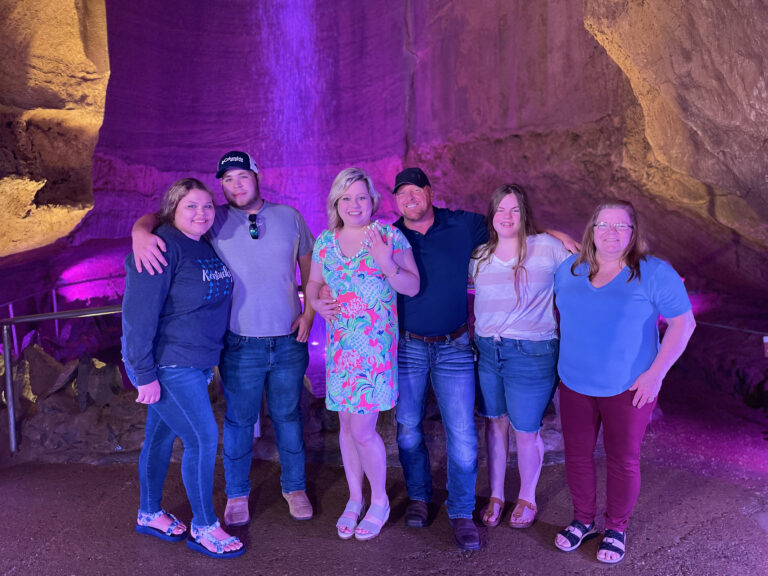 A group of people stand with arms around one another in front of a waterfall in a cave.