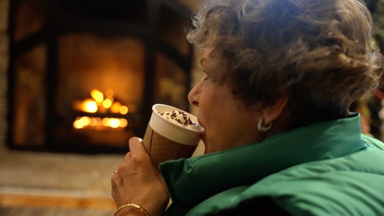 Woman wearing a green vest drinks a seasonal coffee with fireplace in the background.