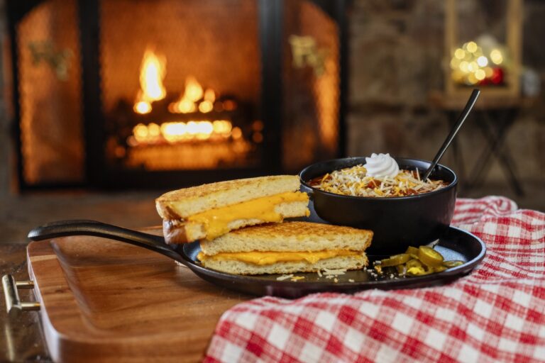 Grilled cheese sandwich and a cast iron skillet with chili in a black bowl topped with cheddar cheese and sour cream. Skillet on a red and white check cloth and wood tray in front of a fire in a fireplace.