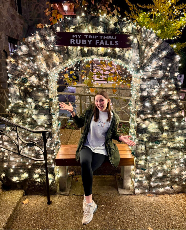 Woman sitting on a bench inside a stone arch wrapped with white lights. A sign above the woman reads 