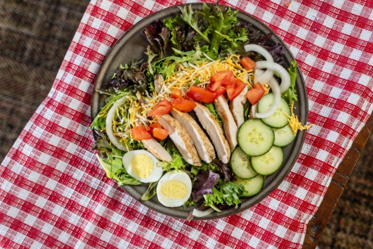 Green salad with sliced grilled chicken, tomatoes, cheese, and cucumbers on a brown plate sitting on a red checked cloth.